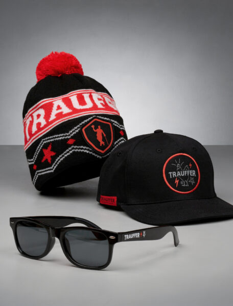 Eine schwarz-rote Strickmütze, eine schwarze Cap und schwarze Sonnenbrillen aus der Trauffer Alpentainer Branded Merch Kollektion, alle mit dem Trauffer-Logo, präsentiert auf einer grauen Fläche vor grauem Hintergrund